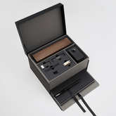 An open black gift box on a light gray background, revealing a wooden pencil box display set, blackwing handheld eraser, blackwing replacement erasers, blackwing point guards, and blackwing notebook clip on the top layer, and a large black spiral bound notebook and medium hardcover notebook with a black pencil in the holster in a lower pull-out drawer with a ribbon string.
