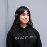 Person leaning against a gray wall wearing black headphones and a black hoodie with 'BLACKWING' wordmark across the front