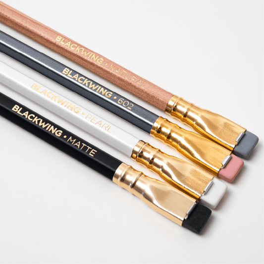 Four different Blackwing pencil types laying on a white background with gold imprint and flat ferrule erasers. Natural, grey, white, and black pencil colors.