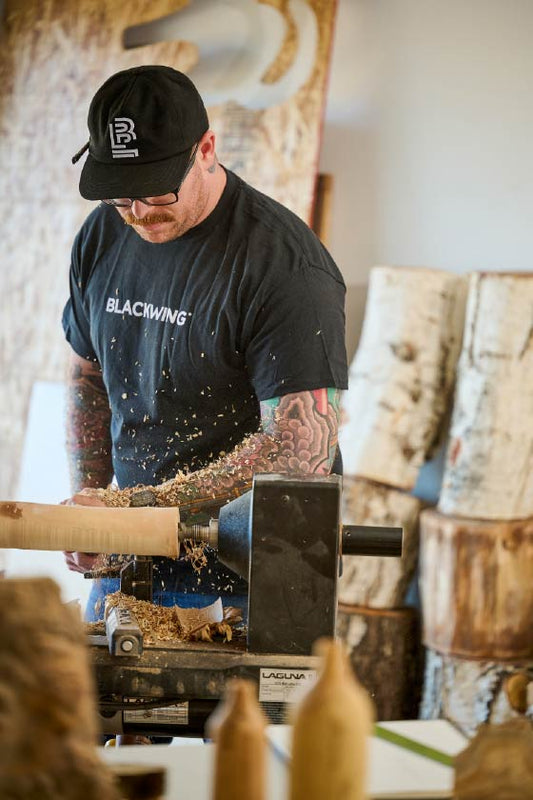 person wearing a black t-shirt with 'BLACKWING' across the front in white and a hat with an embroidered 'B' emblem and a pencil on the side. The person is doing woodwork in a shop.