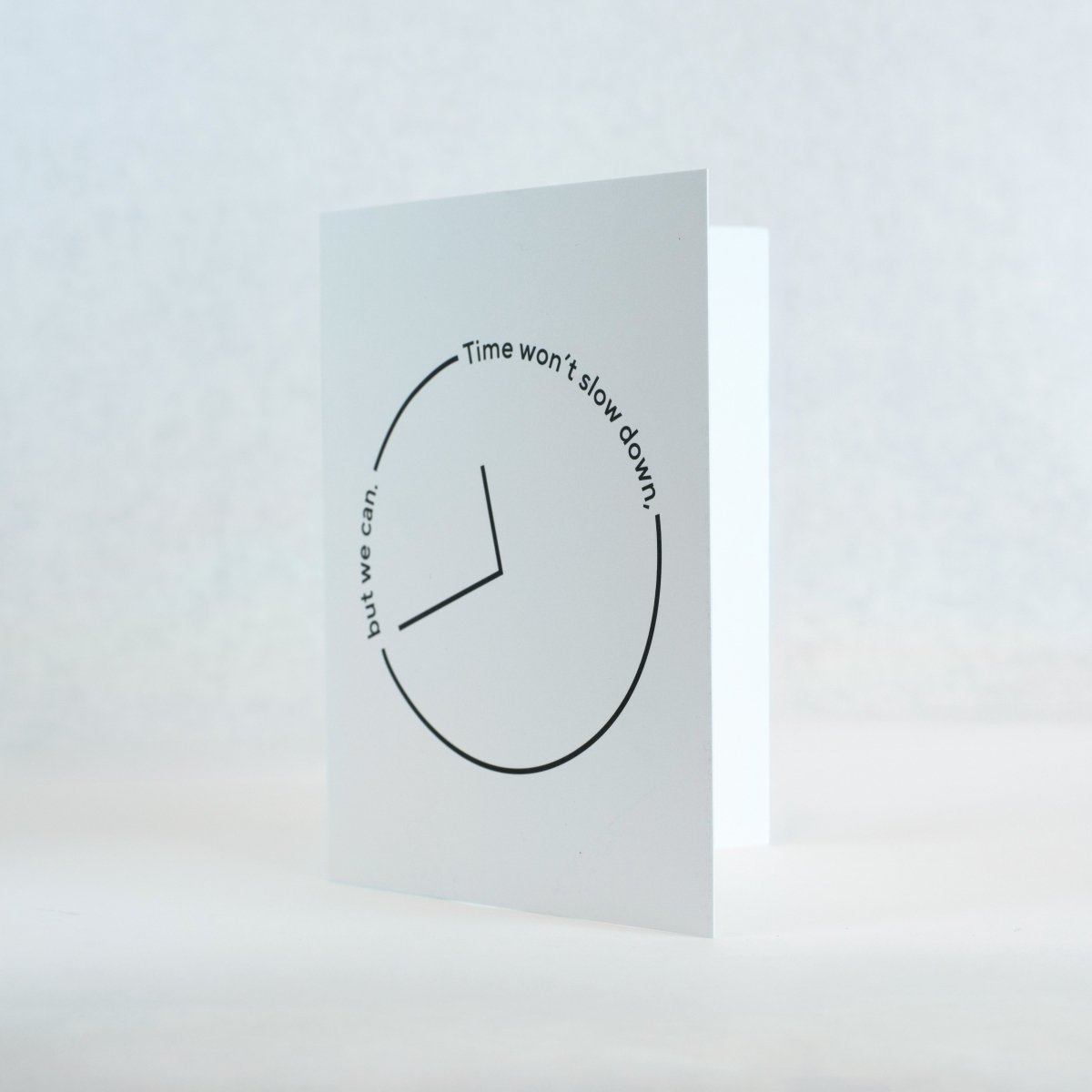white greeting card featuring a black thin outlined clock with the words "time won't slow down, but we can" text along the clock outline.  