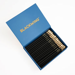 A set of black wood pencils with gold ferrules, black eraser, and gold BLACKWING imprint, neatly arranged inside an open blue presentation box with the brand name BLACKWING printed in gold on the lid, shot on a clean white background.