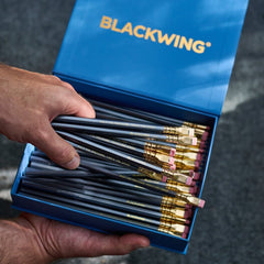 A hand holding an open blue Blackwing pencil box, showing multiple smoky grey pencils with gold ferrules and pink erasers inside. The brand name 'BLACKWING' is printed in gold on the blue lid.