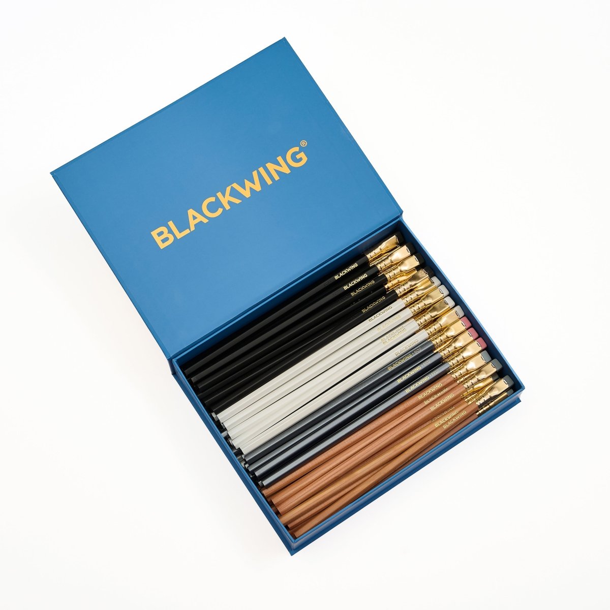 An open blue BLACKWING gift box containing a set of pencils in black, white, gray, and natural wood finishes, with gold ferrules and the BLACKWING logo printed in gold on the lid.