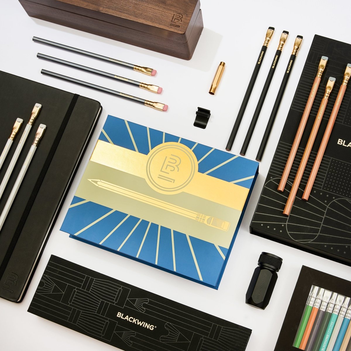 Blackwing stationery arranged on a white background, featuring a blue and gold branded box, black pencils, natural wood pencils, white pencils, silver pencils, a wooden pencil box, black notebook clip, black sharpener, colored pencils, a gold point guard, and a black and gold gift box.