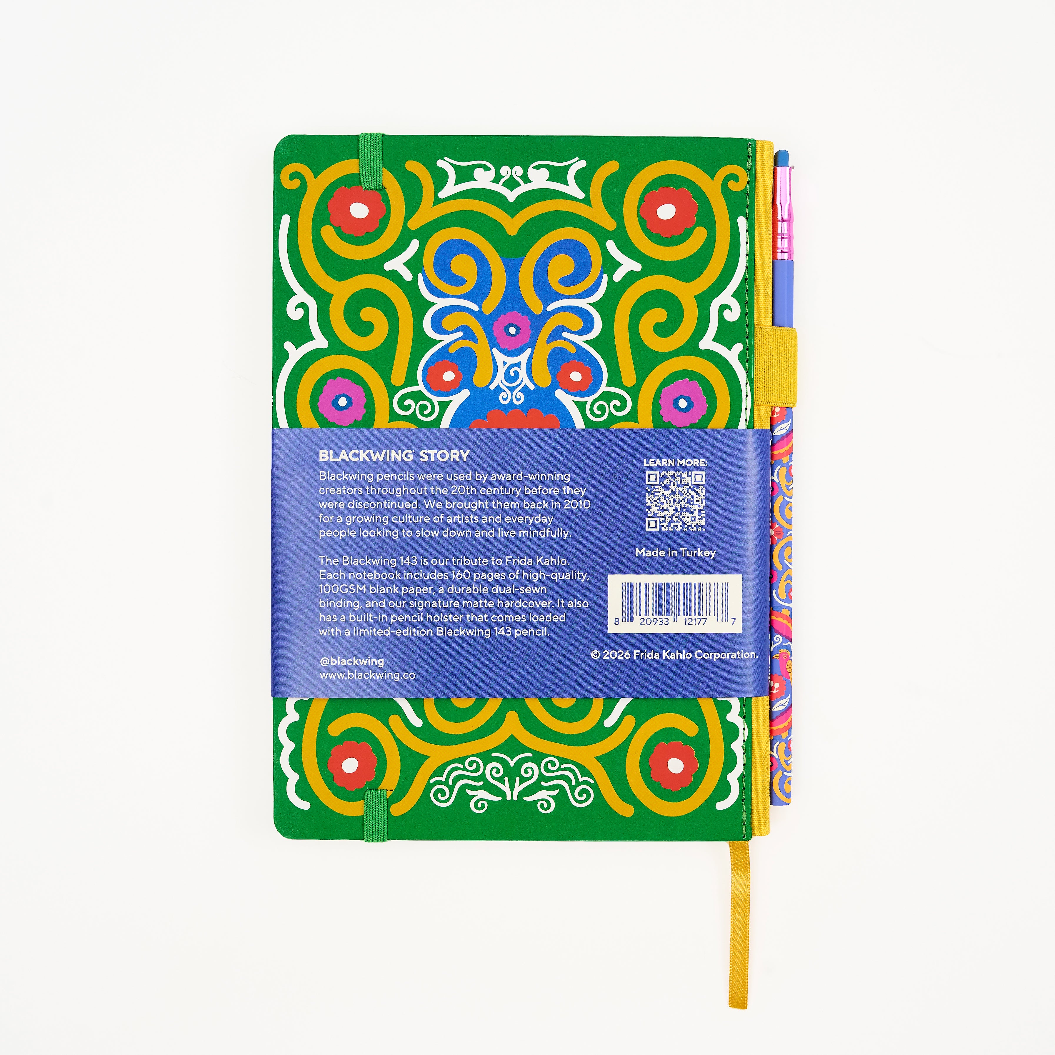 Back of a hardcover notebook featuring a vibrant green cover decorated with a colorful Frida Kahlo art pattern. The notebook is wrapped in a blue paper band with product details, barcode, and a QR code, and is shown with a colorful matching pencil on the right side holster.