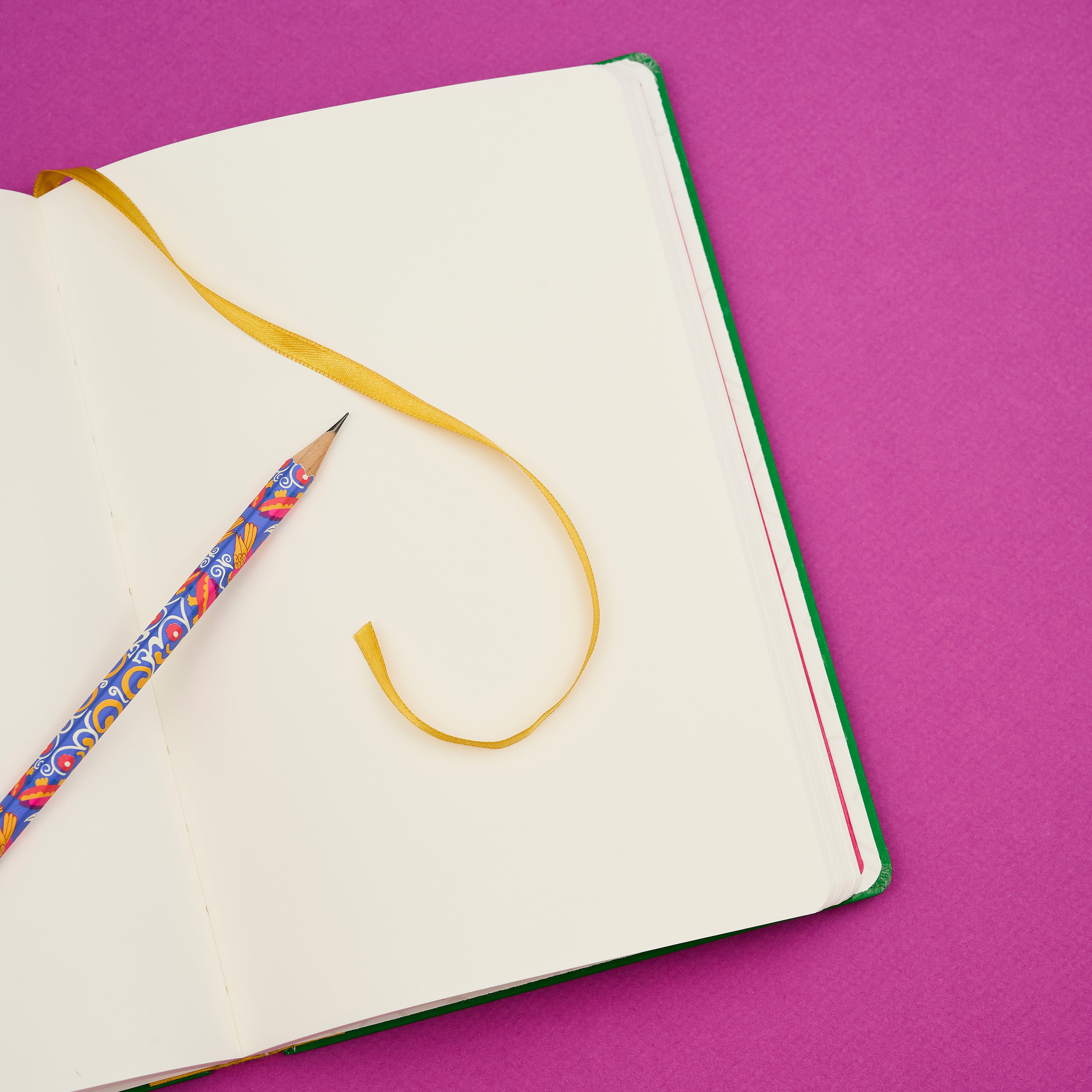An open blank notebook with a yellow ribbon bookmark and a colorful patterned pencil on a solid magenta background.