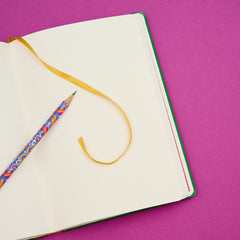 An open blank notebook with a yellow ribbon bookmark and a colorful patterned pencil on a solid magenta background.