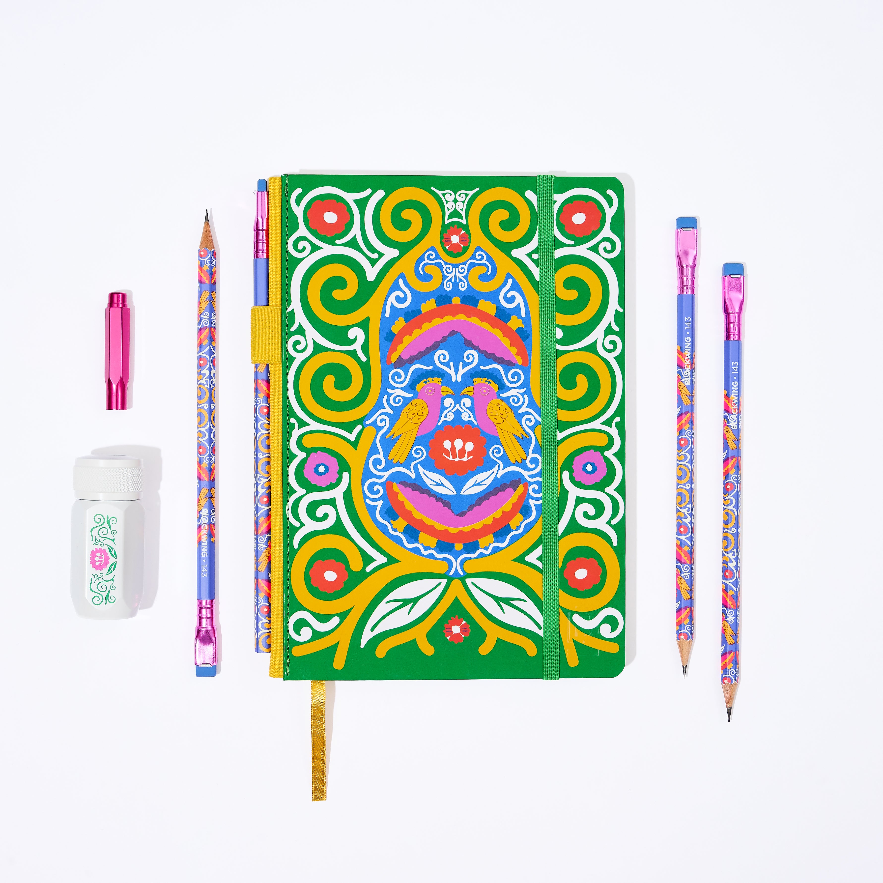 Colorful Frida Kahlo stationery gift set that includes a decorative green, yellow, pink, red, and blue notebook with Frida Kahlo's artwork, several matching patterned pencils, a white sharpener with a floral illustration, and a pink point guard.