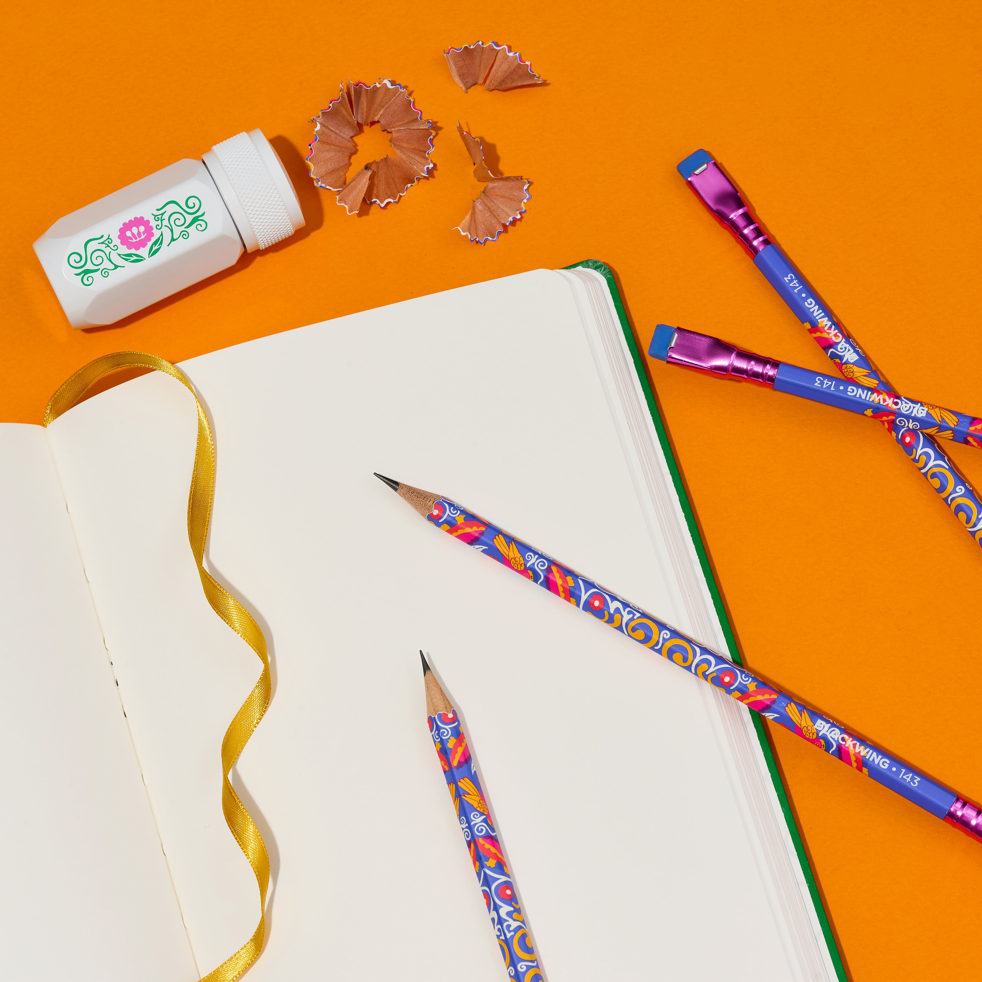 A styled flat-lay of colorful wooden pencils with blue and multi-color barrels, a white sharpener with a pink flower, an open blank notebook, a yellow ribbon, and pencil shavings on a bright orange background.