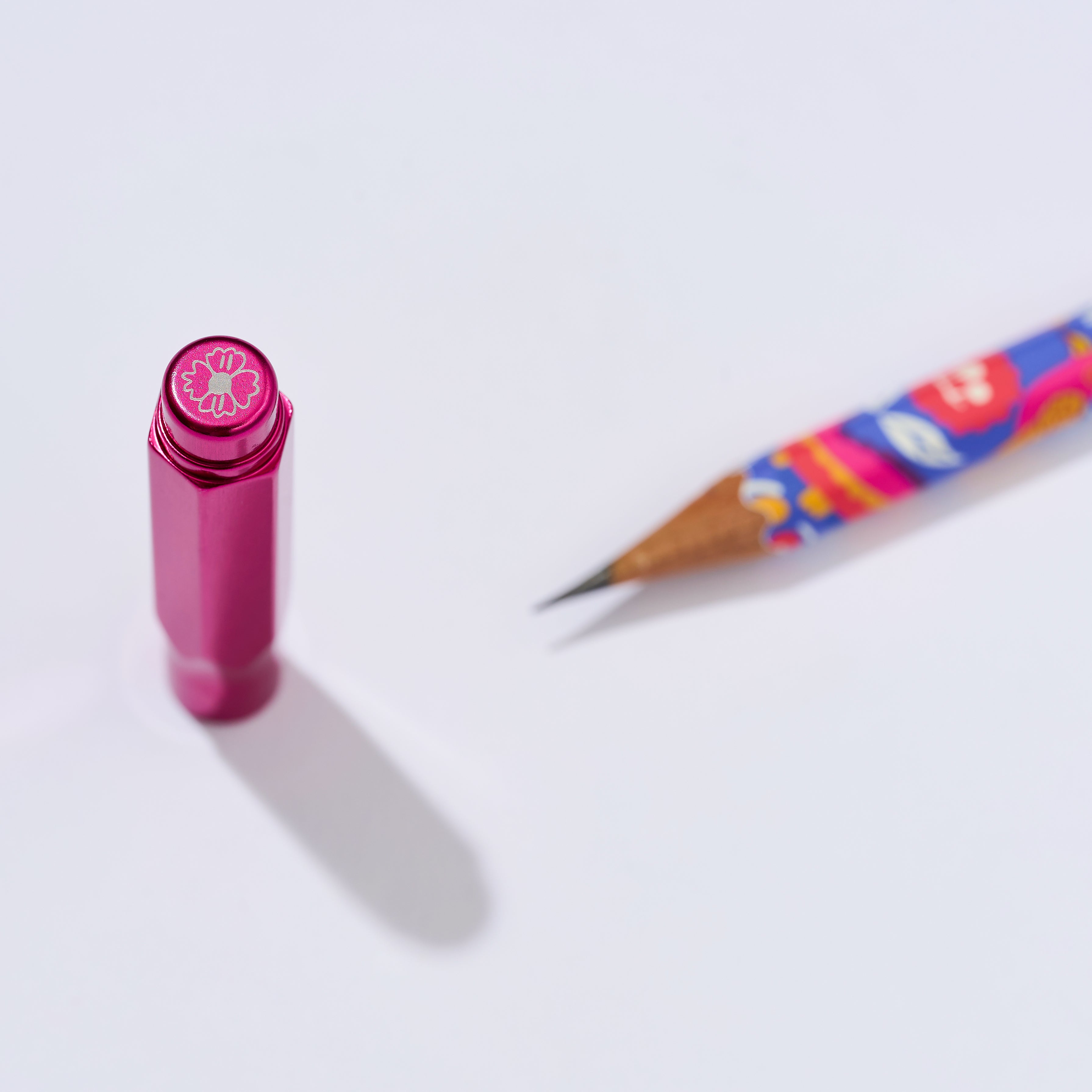 A metallic pink hexagonal point guard with an etched floral emblem on the tip next to a colorful sharpened pencil. 