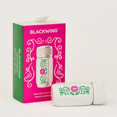 A Blackwing pencil sharpener shown next to its retail box. The sharpener is white with a green and pink floral scroll design, and the box is bright pink and green with the brand name BLACKWING printed in white.