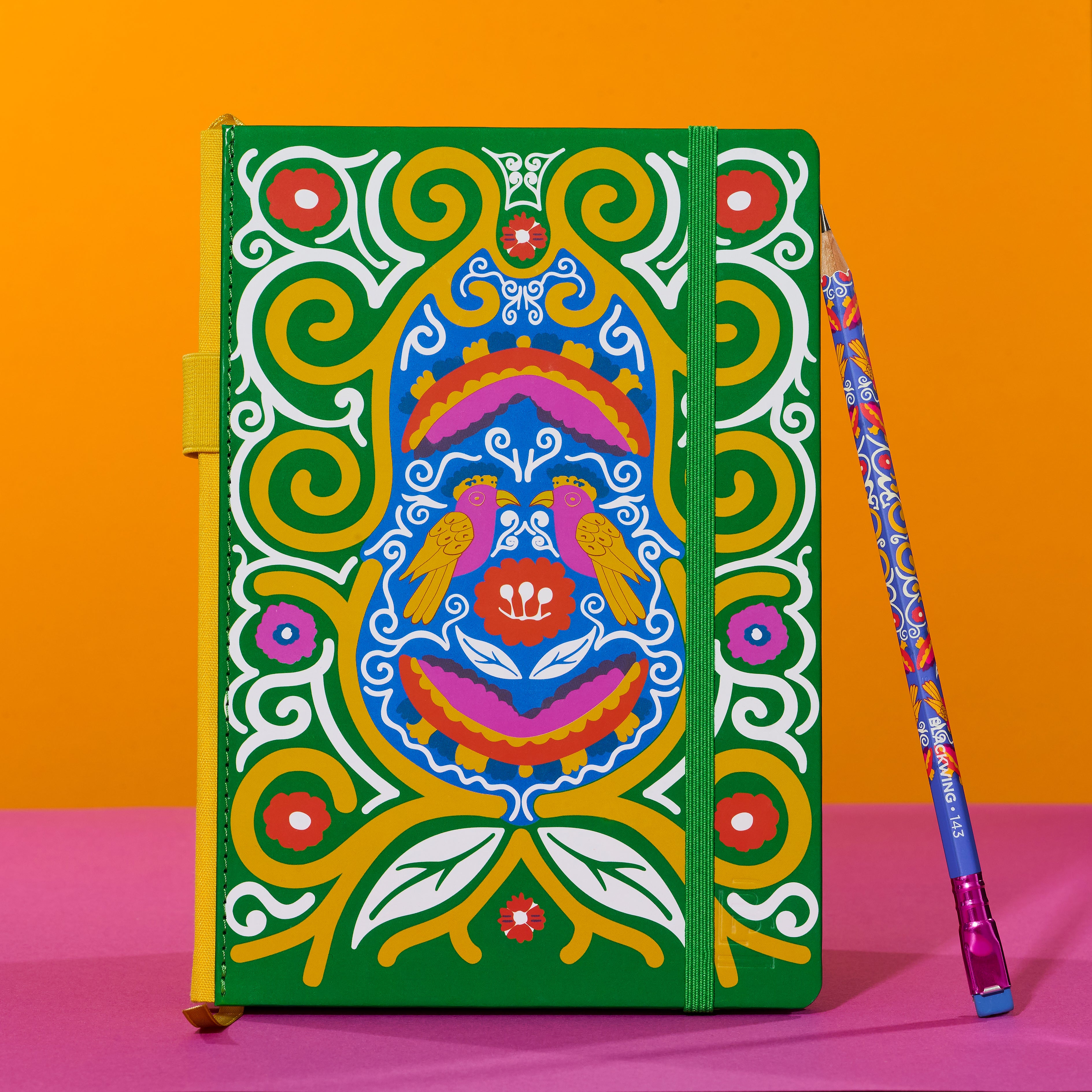 A hardcover notebook with a vibrant, multicolored Frida Kahlo design featuring birds and swirl patterns on a green background, shown next to a matching patterned pencil on a pink and orange background.