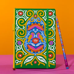 A hardcover notebook with a vibrant, multicolored Frida Kahlo design featuring birds and swirl patterns on a green background, shown next to a matching patterned pencil on a pink and orange background.