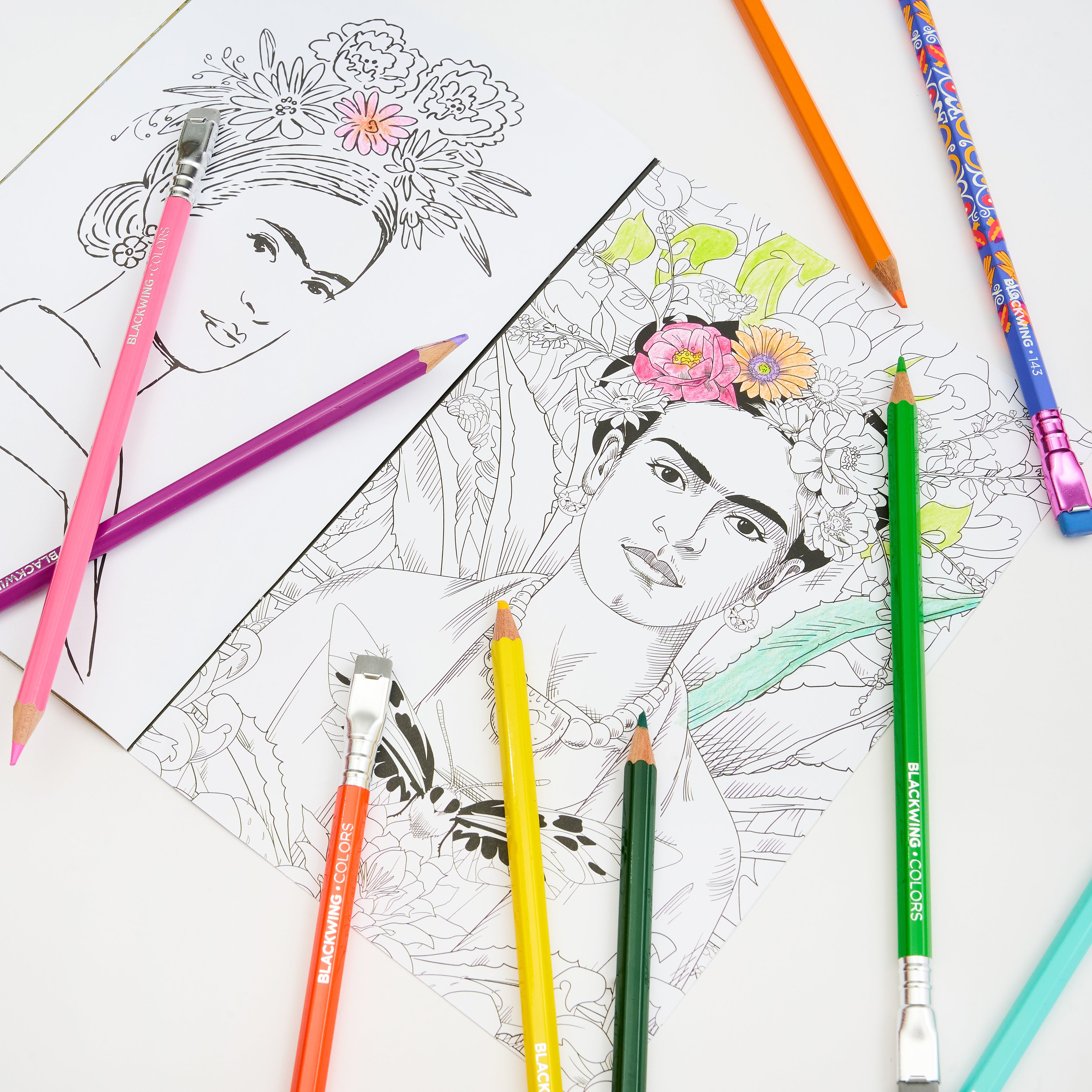 A styled flat-lay of coloring pencils and art supplies on a white surface, including a partially colored portrait drawing of Frida Kahlo with floral details, an uncolored black-and-white version, and several colored pencils in pink, purple, orange, yellow, green, blue, and teal.