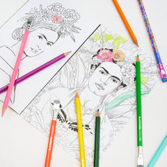 A styled flat-lay of coloring pencils and art supplies on a white surface, including a partially colored portrait drawing of Frida Kahlo with floral details, an uncolored black-and-white version, and several colored pencils in pink, purple, orange, yellow, green, blue, and teal.