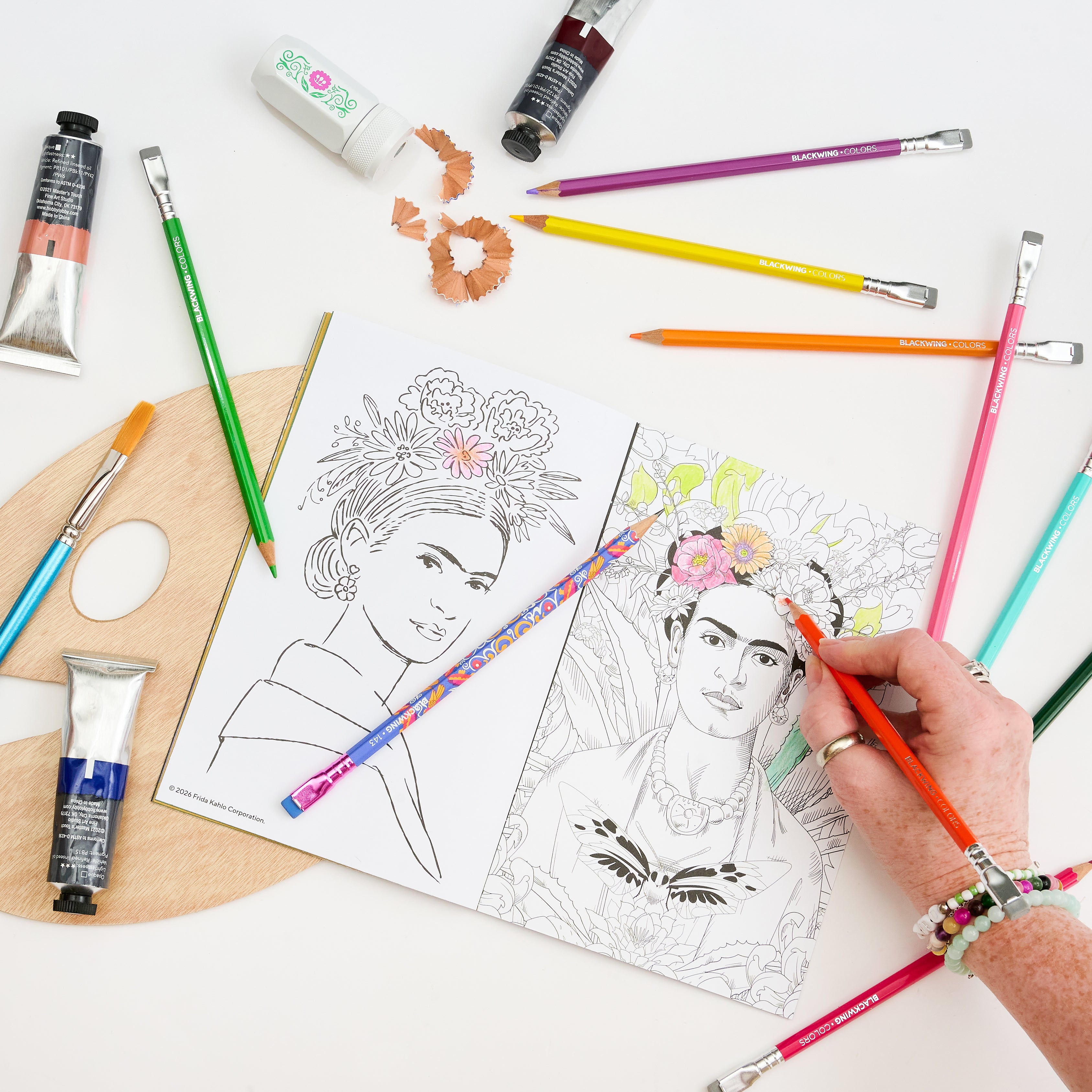 A hand coloring a black-and-white portrait sketch of Frida Kahlo in a coloring book, surrounded by art supplies including paint tubes, colored pencils, a wooden palette, a sharpener, and pencil shavings on a white background.
