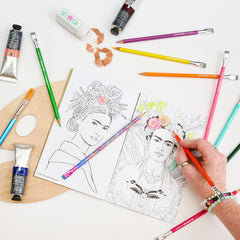 A hand coloring a black-and-white portrait sketch of Frida Kahlo in a coloring book, surrounded by art supplies including paint tubes, colored pencils, a wooden palette, a sharpener, and pencil shavings on a white background.