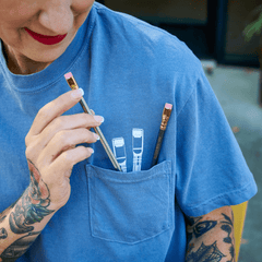 Close-up of a woman wearing a blue t-shirt that has white pencil graphics poking out of the pocket. 2 real Blackwing pencils sit in the pocket.