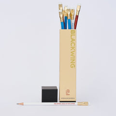 Blackwing pencils in red, white, and blue sticking out of the top of the vertical pencil packaging with one white pencil laying horizontally across the bottom