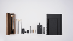 A flat-lay arrangement of stationery and art tools on a clean white background, including a wooden display case with pencils inside, a spiral notebook, a hardcover notebook, a black desktop sharpener, and small pencil accessories.