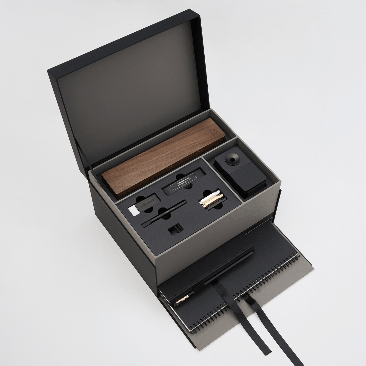 An open black gift box on a light gray background, revealing a wooden pencil box display set, blackwing handheld eraser, blackwing replacement erasers, blackwing point guards, and blackwing notebook clip on the top layer, and a large black spiral bound notebook and medium hardcover notebook with a black pencil in the holster in a lower pull-out drawer with a ribbon string.
