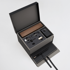 An open black gift box on a light gray background, revealing a wooden pencil box display set, blackwing handheld eraser, blackwing replacement erasers, blackwing point guards, and blackwing notebook clip on the top layer, and a large black spiral bound notebook and medium hardcover notebook with a black pencil in the holster in a lower pull-out drawer with a ribbon string.