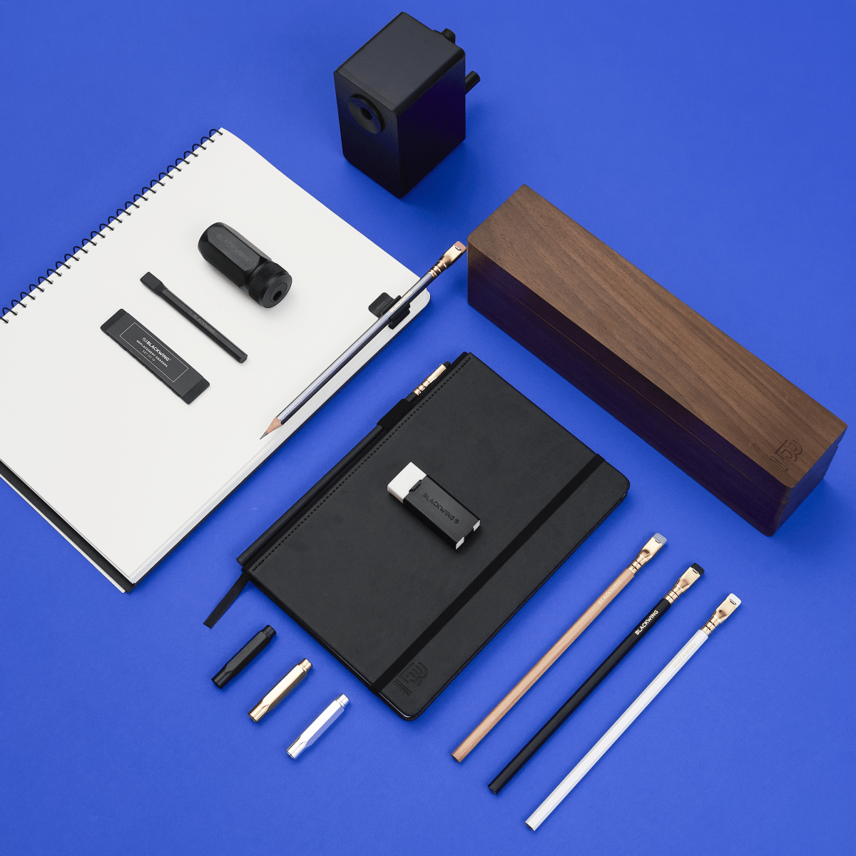 A flat-lay arrangement of blackwing stationery and office accessories on a bright blue background, including a white paper spiral notebook, a black hardcover notebook, several pencils, a black pencil sharpener, and a long rectangular wooden box.