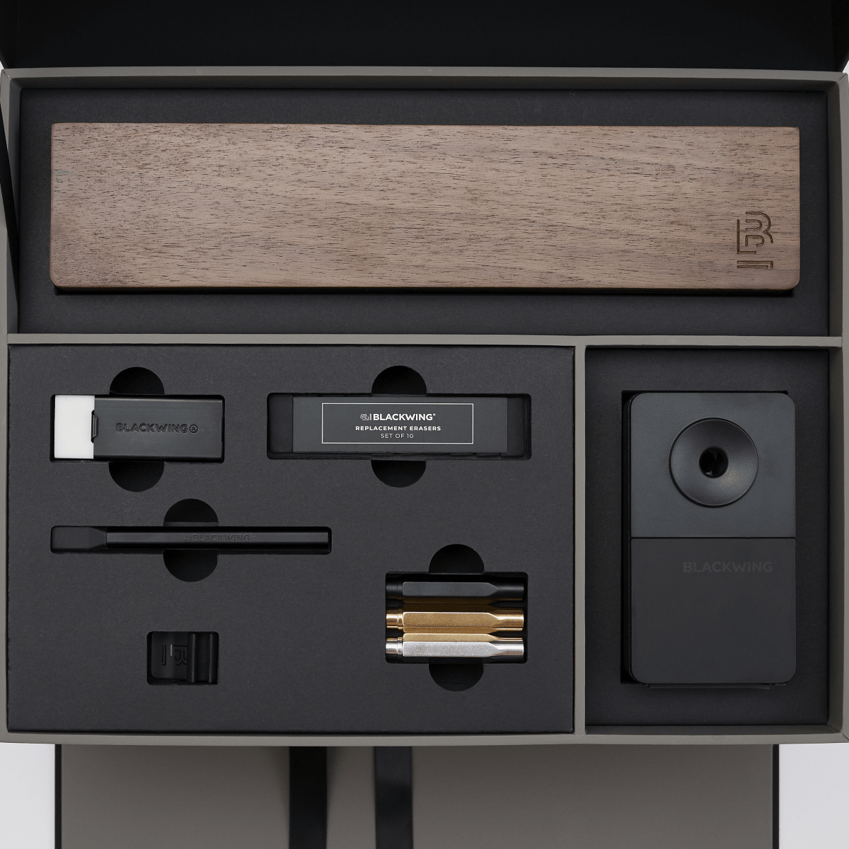 Blackwing stationery items set in a premium presentation box, featuring a wood display case, matte black pencil extender, black and white handheld eraser, black replacement erasers, a black desktop sharpener, a black notebook clip, and silver, gold, and black point guard pencil protectors.