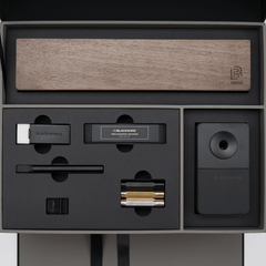 Blackwing stationery items set in a premium presentation box, featuring a wood display case, matte black pencil extender, black and white handheld eraser, black replacement erasers, a black desktop sharpener, a black notebook clip, and silver, gold, and black point guard pencil protectors.