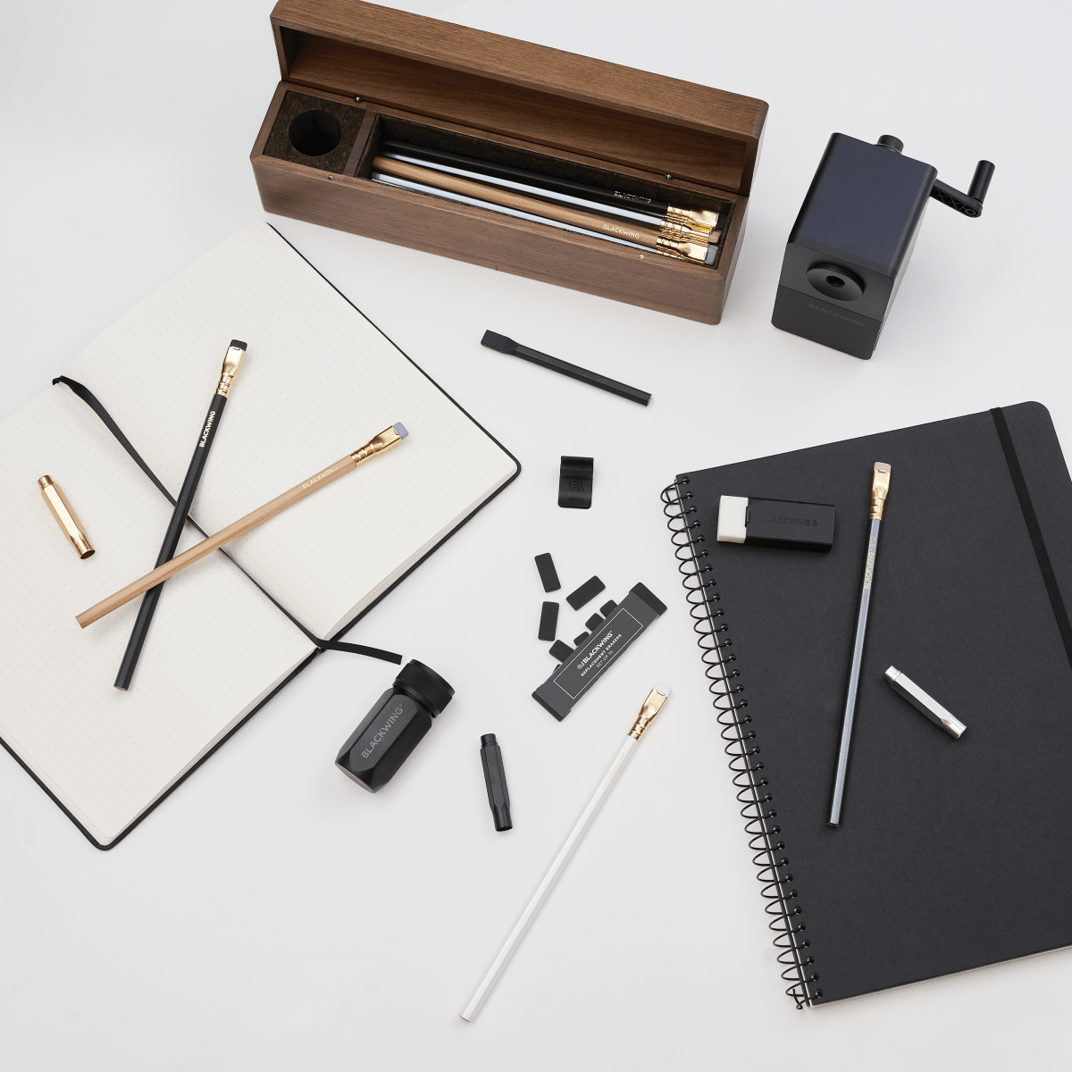 A flat-lay arrangement of stationery and office accessories on a white surface, including a wooden pencil case with pencils inside, a black spiral notebook with a pencil, eraser, and point guard on top, an open notebook with dot-grid paper and two pencils and a point guard on top, a black desktop pencil sharpener, and several black accessories.