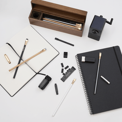 A flat-lay arrangement of stationery and office accessories on a white surface, including a wooden pencil case with pencils inside, a black spiral notebook with a pencil, eraser, and point guard on top, an open notebook with dot-grid paper and two pencils and a point guard on top, a black desktop pencil sharpener, and several black accessories.