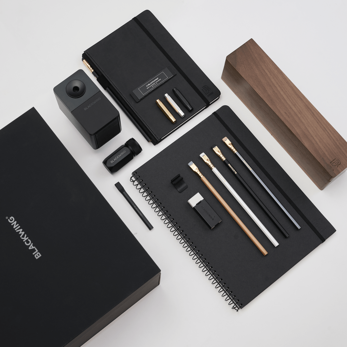 A black stationery and office accessory set arranged on a light gray background, including notebooks, a wood pencil display case, a pencil sharpener, a pencil extender, and other accessories and branded packaging. The visible brand text reads BLACKWING.