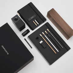 A black stationery and office accessory set arranged on a light gray background, including notebooks, a wood pencil display case, a pencil sharpener, a pencil extender, and other accessories and branded packaging. The visible brand text reads BLACKWING.
