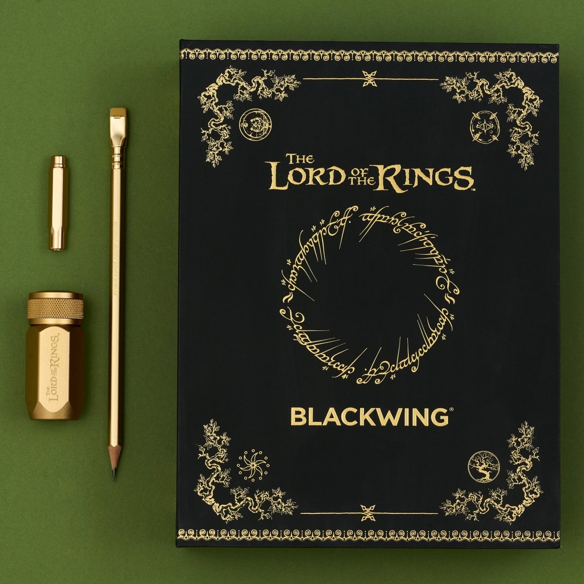 Black box with 'The Lord of the Rings' lettering and gold imprint designs next to a gold pencil, gold point guard, and gold sharpener on a green background