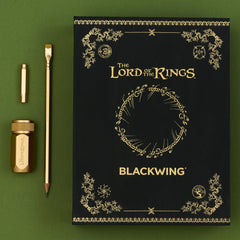 Black box with 'The Lord of the Rings' lettering and gold imprint designs next to a gold pencil, gold point guard, and gold sharpener on a green background