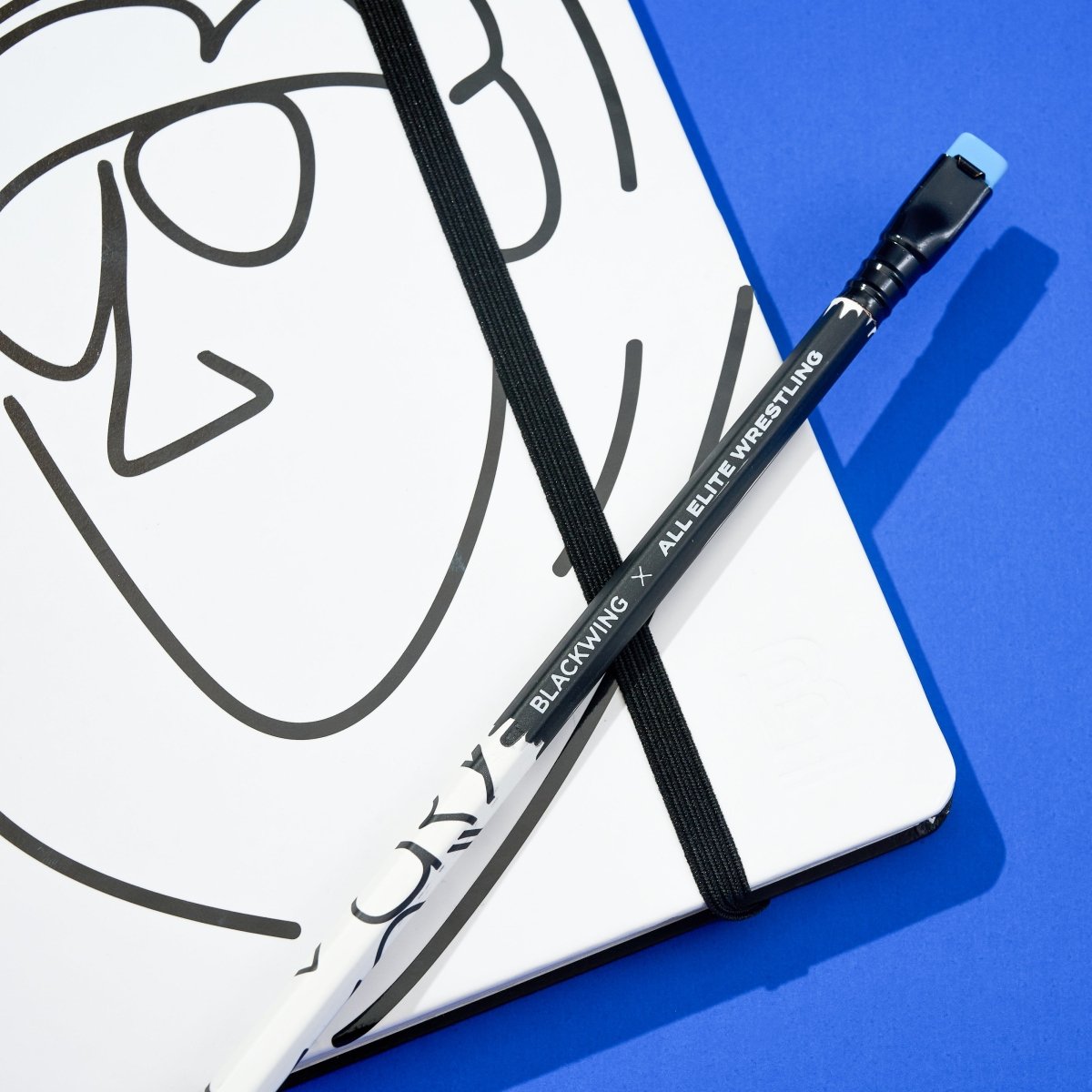 Blackwing x AEW "Freshly Squeezed" Orange Cassidy Notebook with a minimalist black line drawing of a face on a white cover with a matching pencil on top, all on a blue background