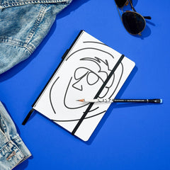 Blackwing x AEW "Freshly Squeezed" Orange Cassidy Notebook with a minimalist black line drawing of a face on a white cover with a matching pencil, all on a blue background with a pair of sunglasses and jean jacket