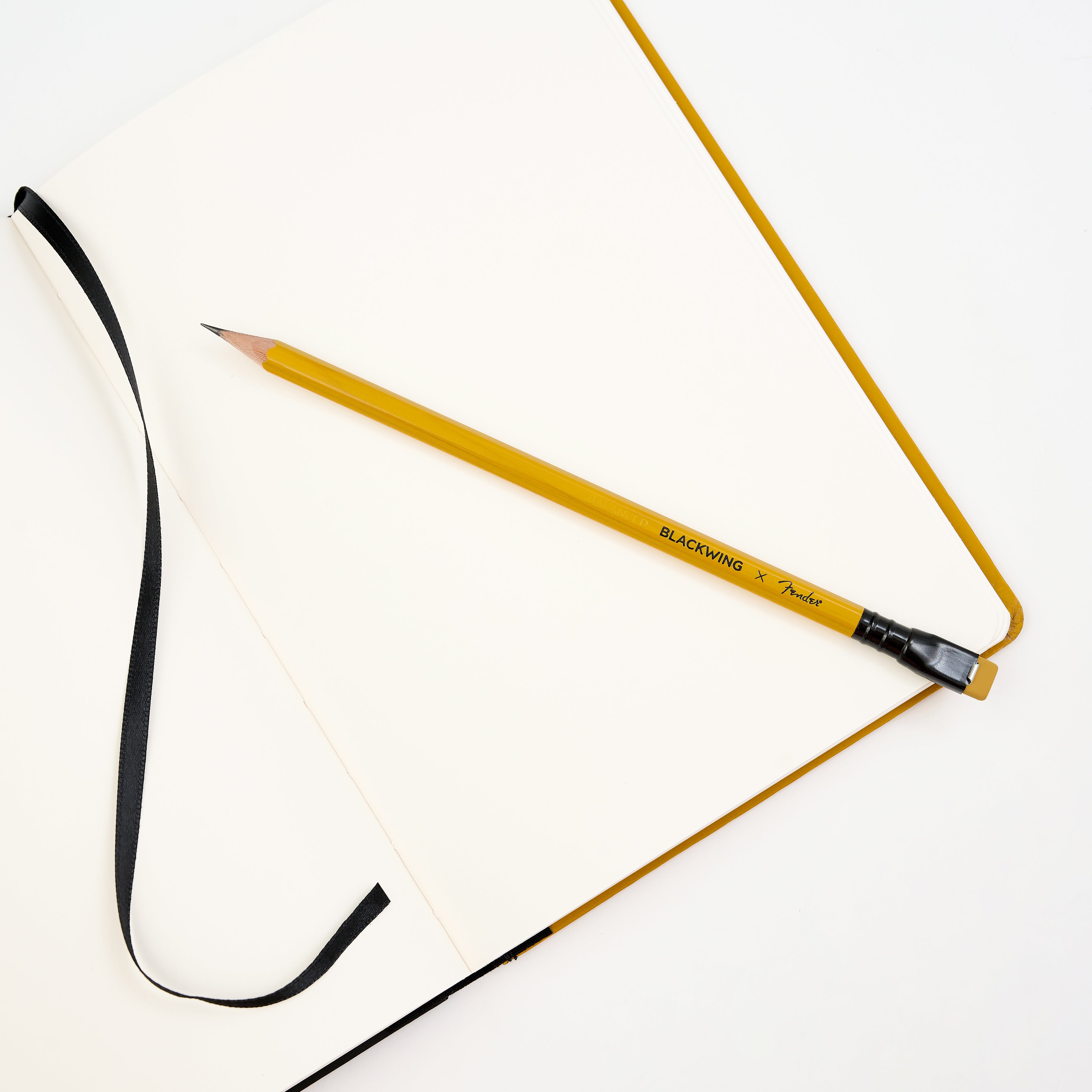 A yellow cedar pencil with a black ferrule and yellow eraser rests diagonally across an open blank notebook with a black ribbon bookmark on a clean white background.