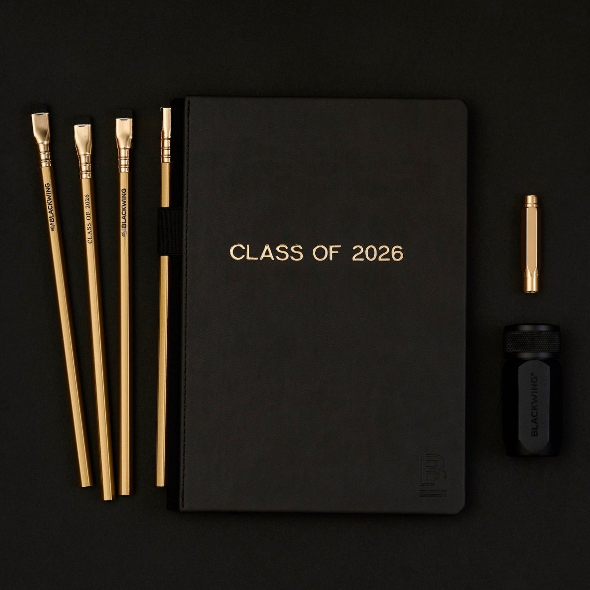 Black notebook with 'Class of 2026' gold foil stamped imprint with 4 gold pencils, a black sharpener, and a gold point guard on a black background