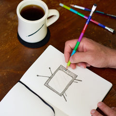 Person drawing a picture of an old TV in a notebook with colorblock pencil, next to a mug of coffee on a wooden table.