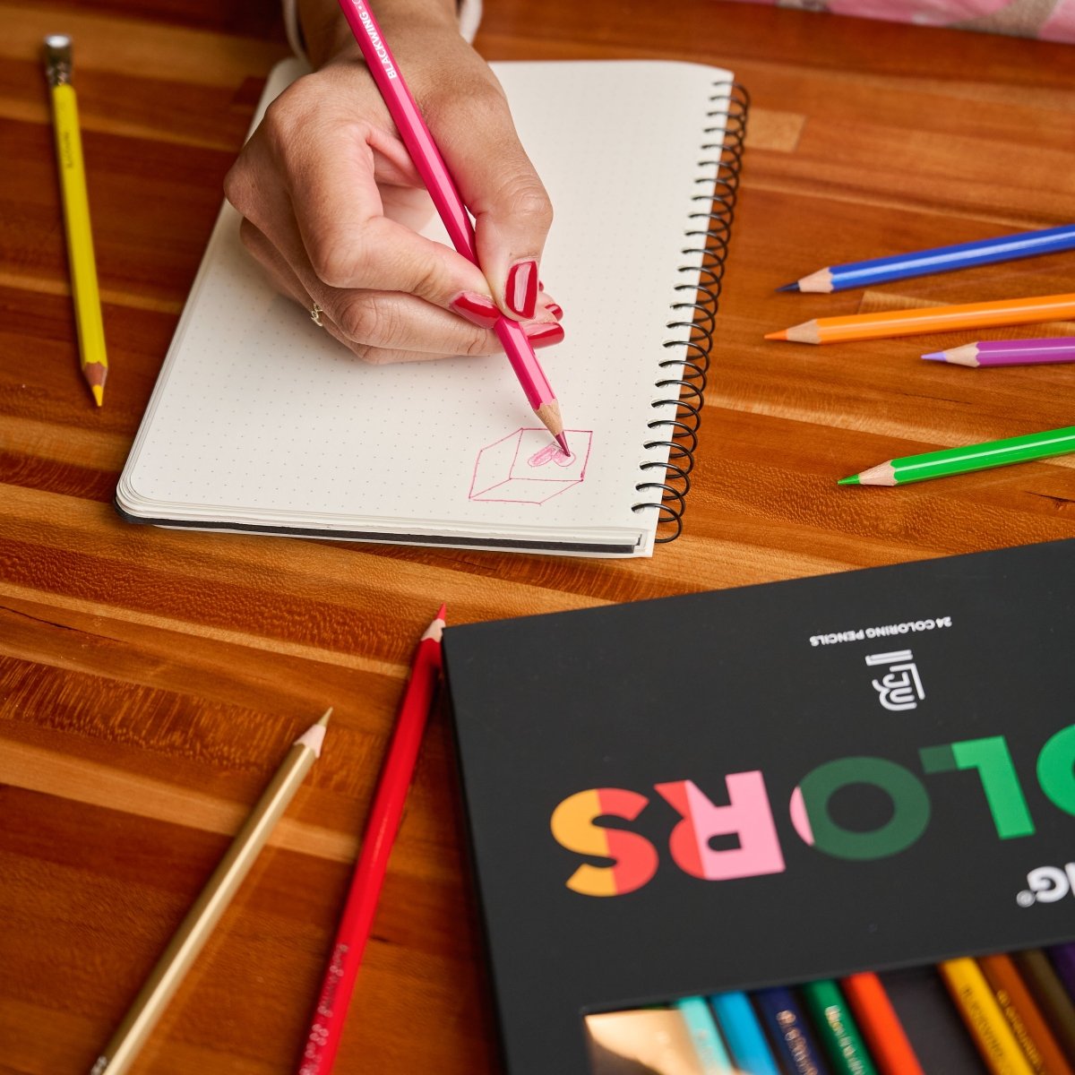 person using a pink colored pencil in a spiral-bound notebook which sits on a wood desk next to other colored pencils