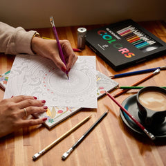 Person coloring a mandala in a coloring book with various colored pencils on top of a wood desk with a sharpener and coffee 
