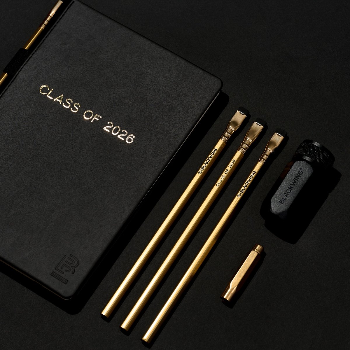 Black notebook with 'Class of 2026' gold foil stamped imprint across the front with a gold pencil in the holster (biinding), sitting next to 3 more gold pencils, a black sharpener, and a gold point guard on a black background
