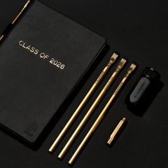 Black notebook with 'Class of 2026' gold foil stamped imprint across the front with a gold pencil in the holster (biinding), sitting next to 3 more gold pencils, a black sharpener, and a gold point guard on a black background