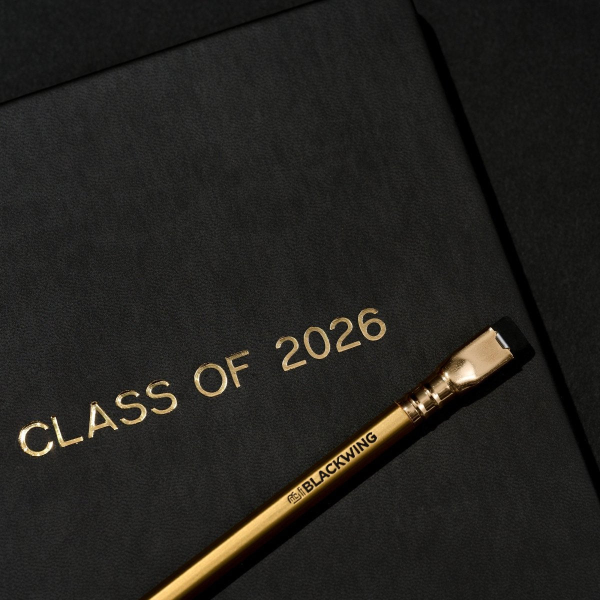 'Class of 2026' black notebook and a gold pencil on a black background