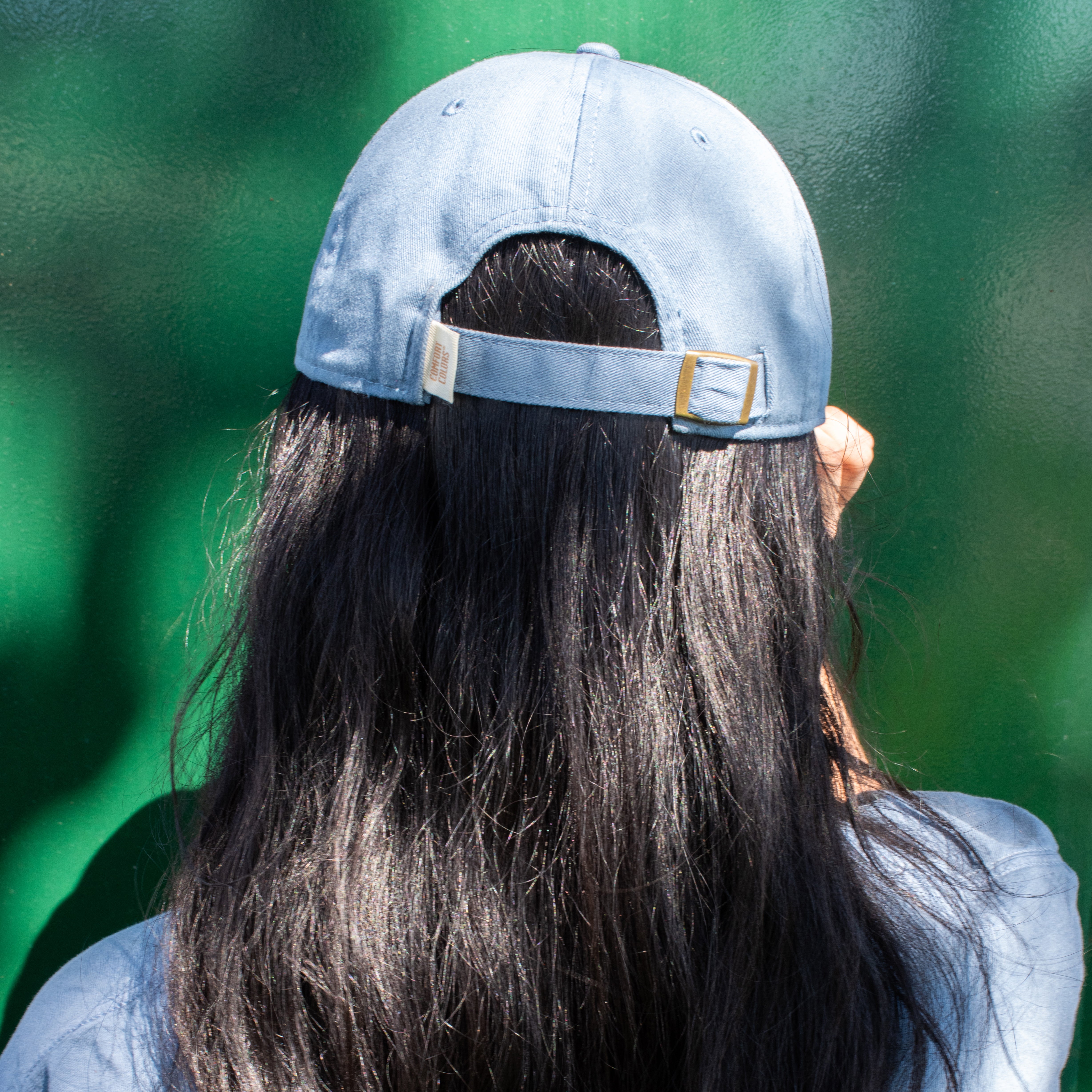 back of a woman's head sporting a blue cotton dad hat and blue t-shirt against a green background