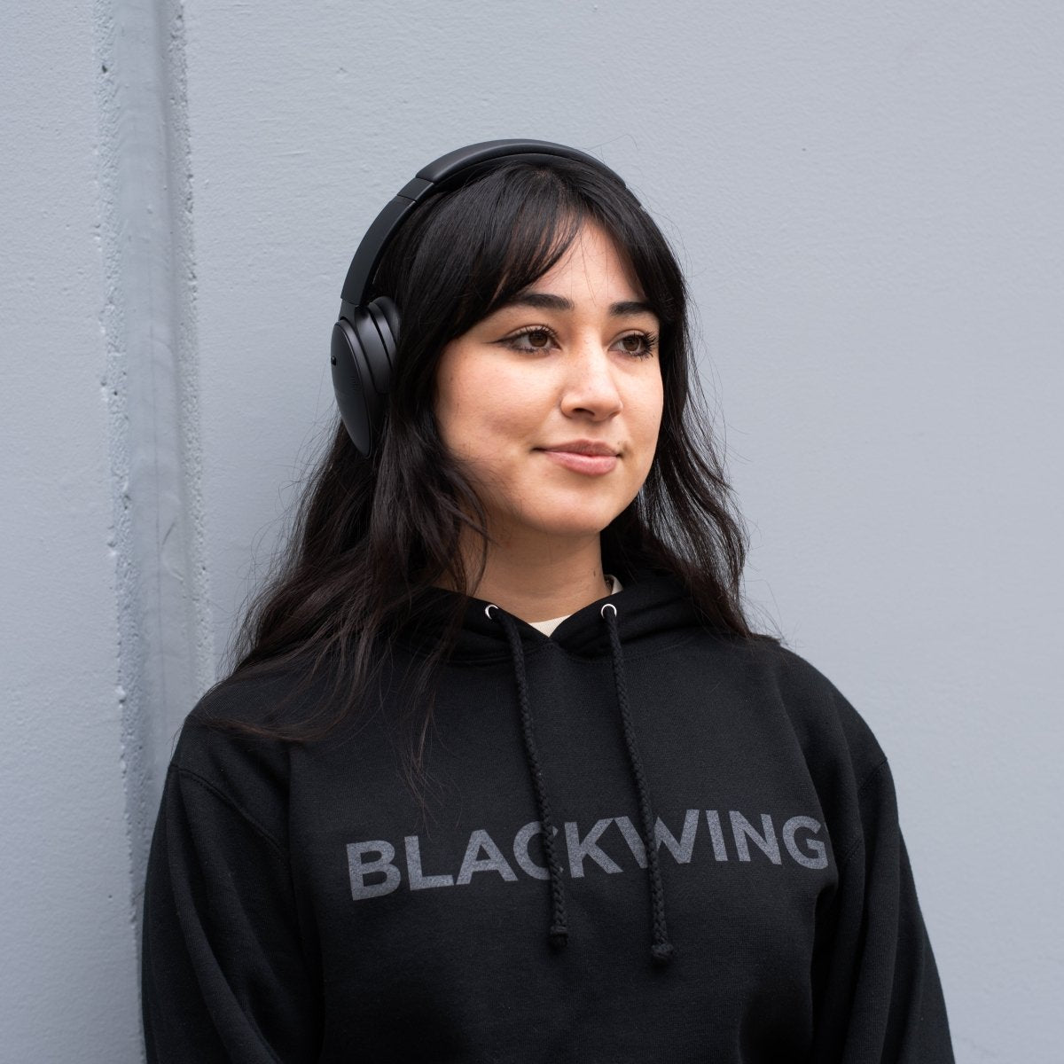 Person leaning against a gray wall wearing black headphones and a black hoodie with 'BLACKWING' wordmark across the front