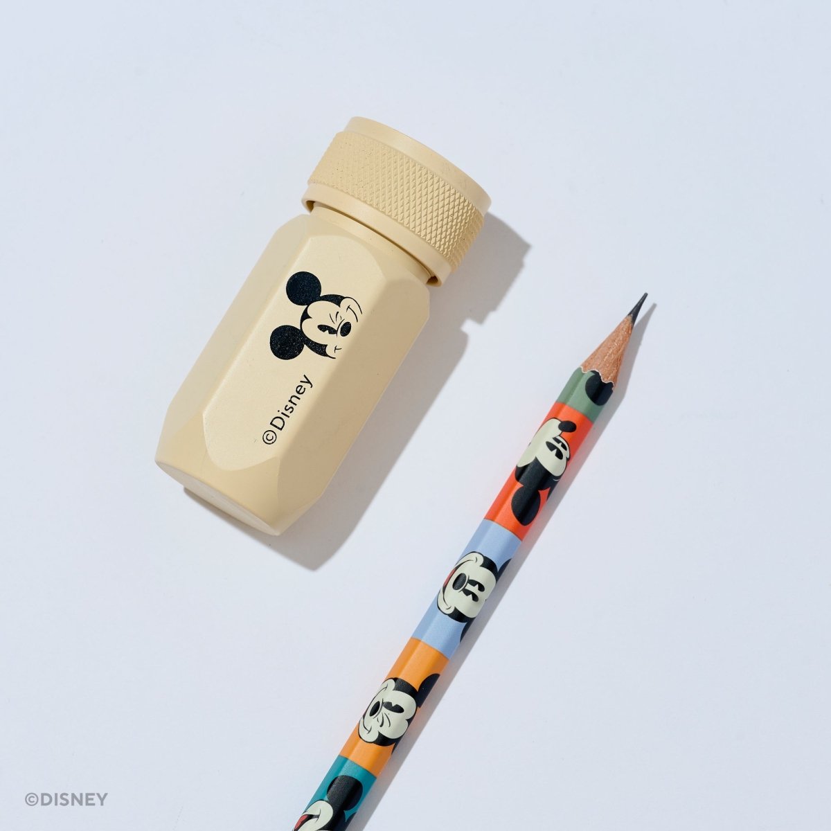 A Disney pencil and pencil sharpener set on a light gray background. The sharpener is a light beige aluminum hexagonal tool with a black Mickey Mouse graphic and the Disney name. The pencil barrel features various Mickey Mouse face graphics in different colorblocks.