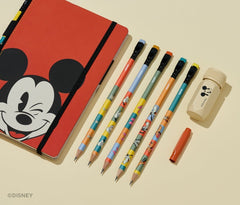 A Disney stationery set arranged on a warm cream background, including a red notebook with a Mickey Mouse face, five colorful pencils, a red pencil guard, and a beige sharpener with a Mickey Mouse head print.